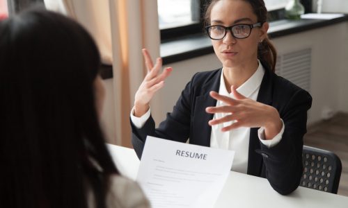 Confident millennial female applicant in glasses talking at job interview answering questions, vacancy candidate making first impression concept and introducing herself speaking to hr holding resume